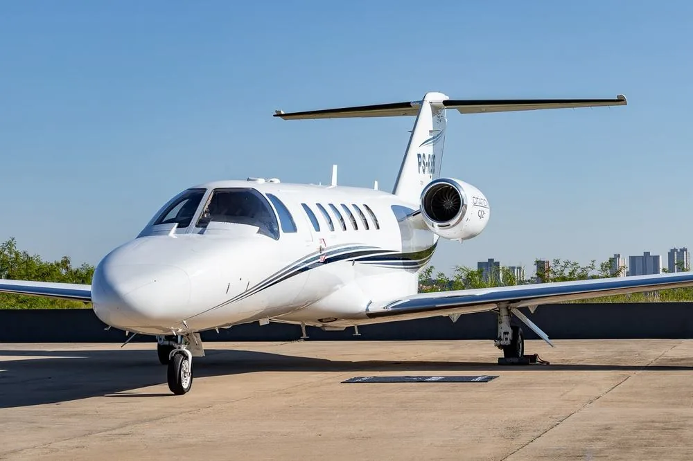 Citation CJ2+ branco com faixa azul e prata, estacionado no pátio sob céu limpo, com a silhueta da cidade ao fundo.