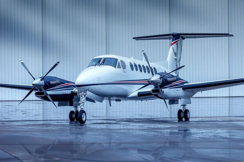 King Air B350 branco com faixas azul e vermelha, em vista frontal de três quartos, estacionado em frente a um hangar.